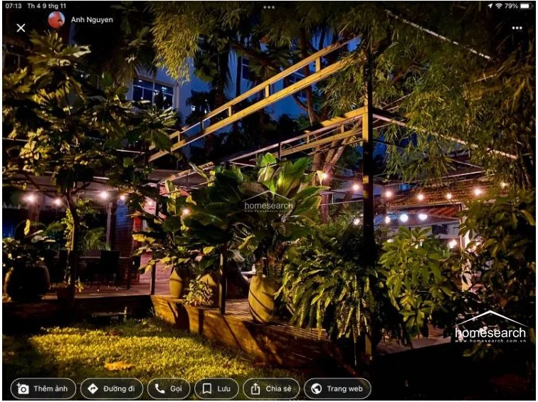 This image captures a lush, atmospheric outdoor terrace at night, illuminated by warm string lighting that creates a cozy, inviting ambiance. The space features a wooden deck surrounded by mature tropical greenery and potted plants, offering a serene retreat from the adjacent building.