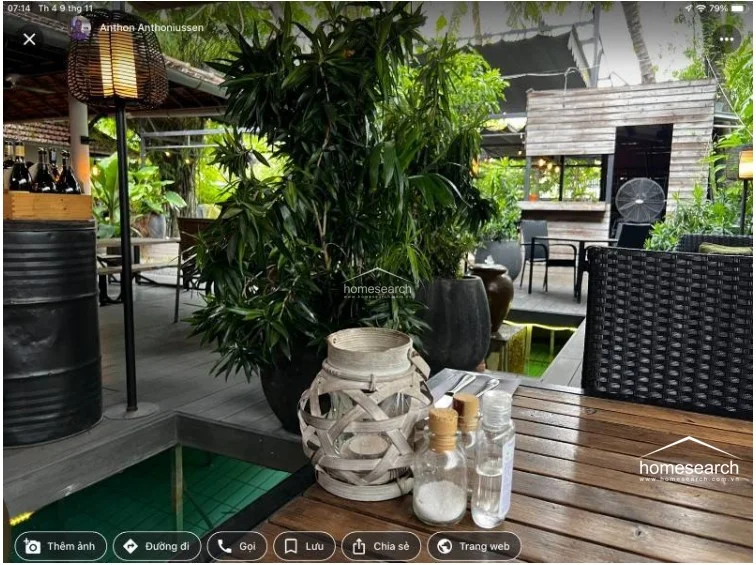 This close-up shot of the outdoor dining area emphasizes a relaxed, rustic aesthetic with a wooden table, woven lantern lighting, and industrial-style barrel accents. The space is thoughtfully decorated with lush potted plants, creating an intimate setting that seamlessly blends indoor comfort with the natural environment.