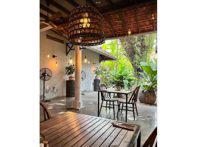 This covered patio area features a rustic, open-concept design with tiled flooring, a prominent central column, and exposed wooden ceiling beams. The space is furnished with simple dining sets and floor fans, while warm wall-mounted lighting and large potted plants enhance the relaxed, tropical atmosphere.