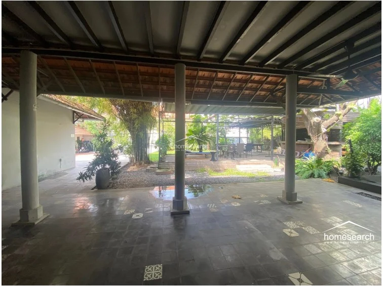 This covered patio serves as an expansive outdoor living area, featuring dark patterned stone flooring and multiple structural columns supporting the roof. The space is open-air, allowing for a natural flow into the surrounding garden and providing a shaded retreat from the sun.