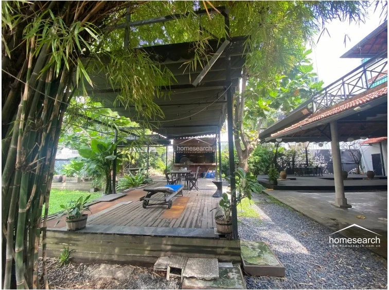 This perspective showcases the functional outdoor living space, which includes a retractable canopy over the wooden deck and a dedicated bar or food preparation area in the background. The transition between the wooden decking, gravel pathways, and green lawn creates a well-defined flow, suitable for outdoor entertaining or leisure.