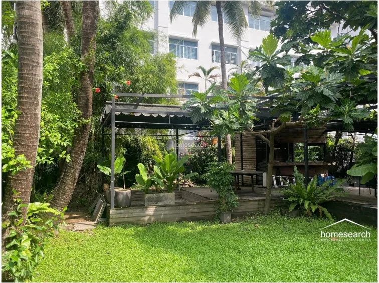 This outdoor space features a raised wooden deck covered by a black metal pergola, creating an ideal area for al fresco dining or relaxation. The garden is lush with mature trees, potted plants, and a well-maintained lawn, offering a serene and private atmosphere.