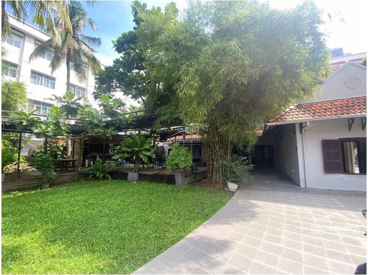This daytime view highlights the property's expansive outdoor layout, featuring a mix of a manicured lawn, a paved driveway, and a raised wooden deck area. The architecture includes a classic structure with a tiled roof, while mature bamboo and trees provide natural shade and privacy to the open-air living space.