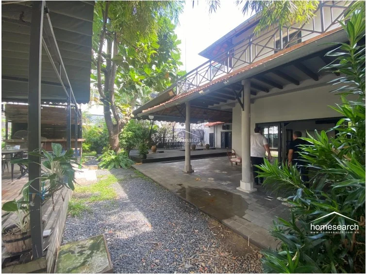 This view shows a spacious, covered patio area with dark stone flooring that seamlessly connects the indoor living space to the garden. The architectural design includes structural columns and a traditional tiled roof, providing a sheltered transition zone between the interior and the backyard.