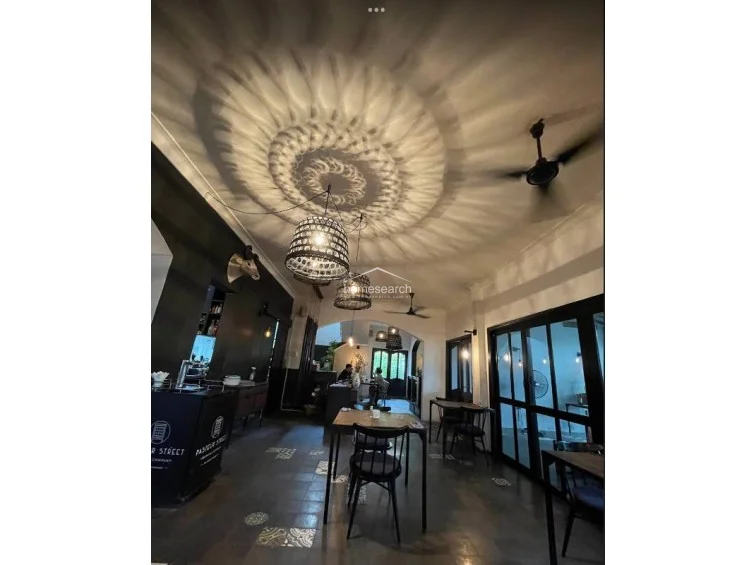 This dining area showcases an intimate, atmospheric design with warm, patterned light projections on the ceiling cast by woven pendant fixtures. The space is furnished with dark wooden tables and chairs, maintaining the cohesive black and white color scheme seen throughout the property. Large glass-paneled doors offer a seamless connection to the outdoor garden area, enhancing the flow of the room.