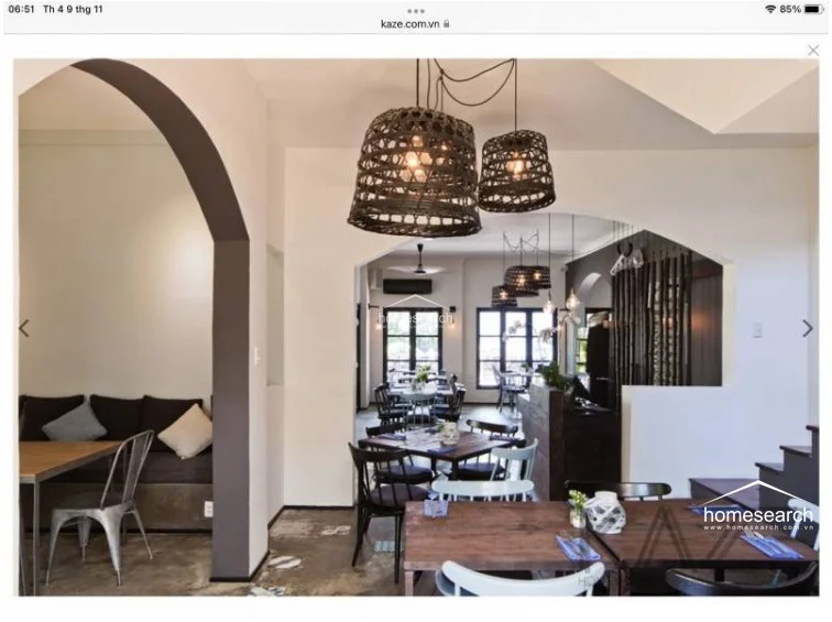 This inviting dining space features a comfortable built-in bench seat with neutral cushions, positioned under a classic architectural archway. The room is illuminated by a cluster of decorative woven pendant lights that create a soft, ambient glow over the wooden tables. The flooring consists of dark, textured stone, and the overall maintenance level is high, reflecting a sophisticated and cozy design.