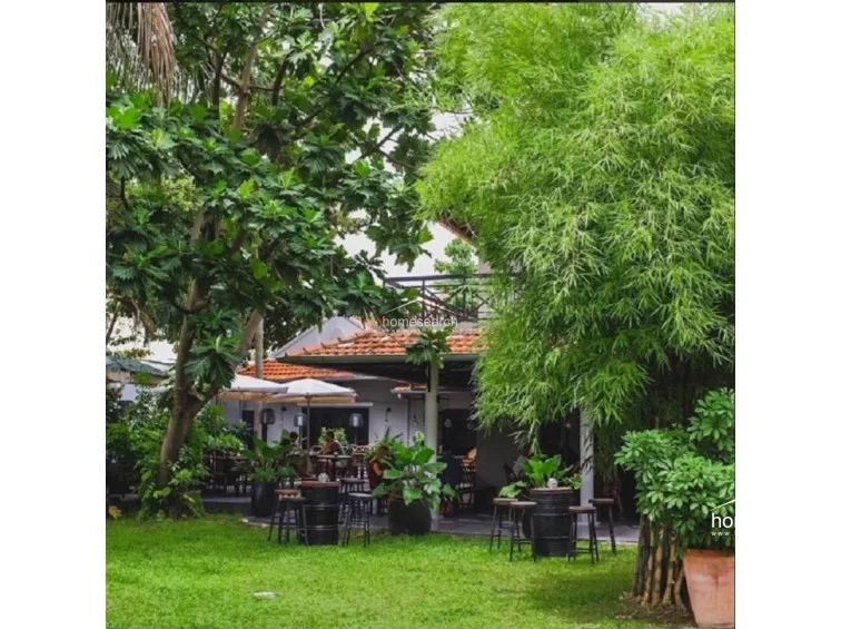 This lush outdoor garden area provides a serene, natural setting with mature trees, bamboo, and vibrant greenery surrounding a paved patio. The space is furnished with casual, industrial-style high-top tables made from repurposed barrels, perfect for an outdoor lounge or dining experience. The area is well-maintained and offers a peaceful, private retreat from the main building.