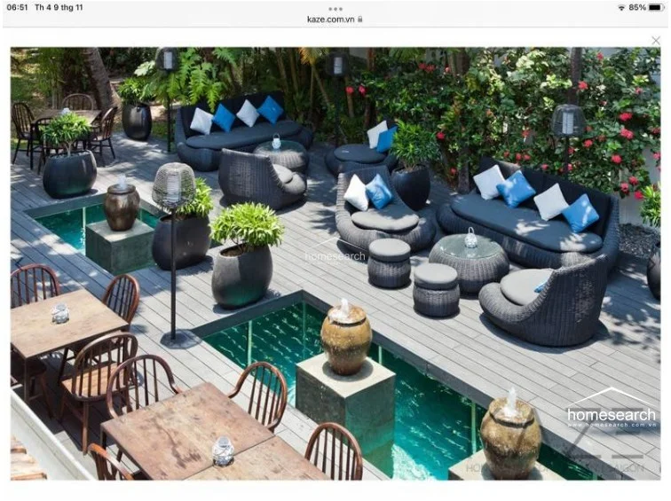 This expansive outdoor terrace features high-quality dark composite decking and is centered around a unique, sunken water feature with decorative stone fountains. The area is furnished with comfortable, modern outdoor wicker lounge sets with plush cushions, creating a luxurious and relaxing atmosphere. The space is beautifully landscaped with potted plants and is ideal for hosting, blending architectural elegance with natural elements.