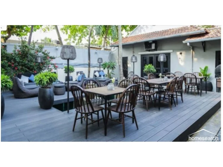 This outdoor dining and lounge area features a spacious wooden deck that seamlessly connects to the interior through large glass-paneled doors. The space is well-furnished with dark wicker seating and wooden dining sets, complemented by modern floor lamps and lush potted greenery that enhances the relaxed, airy atmosphere.