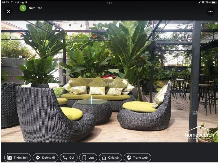 This cozy outdoor seating nook is nestled within a lush garden setting, utilizing comfortable wicker lounge chairs with vibrant lime-green cushions. The area is defined by a wooden deck and a black metal pergola structure, creating an intimate and shaded retreat perfect for lounging.