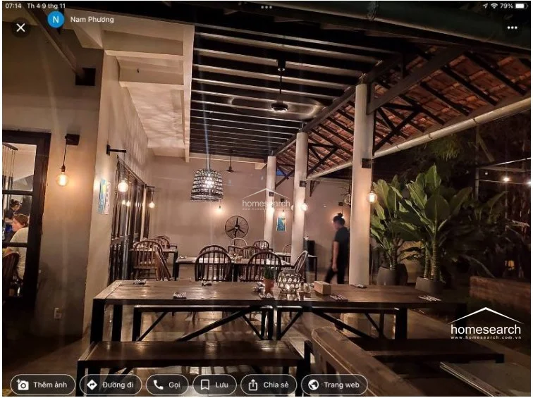 This interior-exterior dining space showcases a rustic-industrial aesthetic with exposed dark wooden ceiling beams and clean, white-painted columns. The layout is open and airy, featuring long wooden tables and benches that provide a communal dining experience, all illuminated by stylish hanging pendant lights.