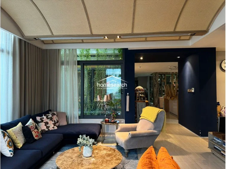 This angle emphasizes the connection between the indoor living space and the outdoor greenery, visible through expansive glass sliding doors draped in floor-length beige curtains. The room's design is cohesive, blending warm textures like the rattan ceiling with contemporary finishes to create an inviting, well-curated living environment.