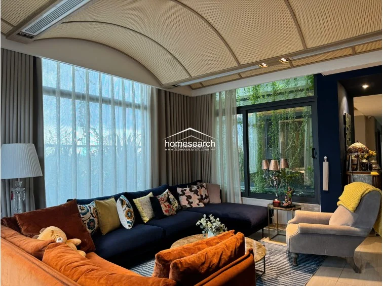 This sophisticated living room features a striking curved, textured ceiling and floor-to-ceiling windows that offer a lush, green view. The space is anchored by a deep navy blue sectional and a contrasting burnt-orange sofa, set against a backdrop of neutral walls and wood-look flooring.
