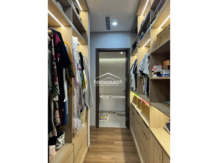 This organized walk-in closet features floor-to-ceiling light wood cabinetry with integrated LED lighting and a mix of hanging rods and open shelving. The layout flows directly into an en-suite bathroom, which is visible through the doorway and contains a clean, white soaking tub.