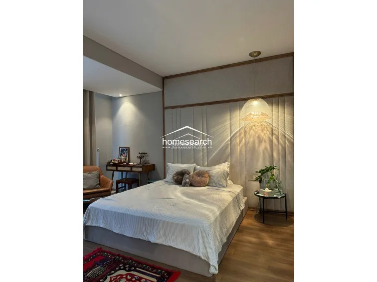 This serene bedroom is defined by a large, floor-to-ceiling upholstered headboard with vertical paneling and a warm, ambient pendant light. The room includes a dedicated vanity area, wood flooring, and large windows draped in neutral curtains that provide ample natural light.