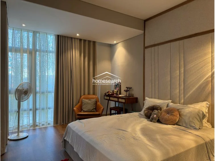 This bedroom layout emphasizes a seamless transition between the sleeping area and the adjacent dressing room and bathroom. The space maintains a cohesive, modern aesthetic with wood flooring, soft grey walls, and a glass-partitioned view into the bathroom’s soaking tub.