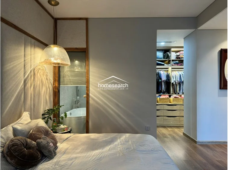 This bedroom view highlights the elegant interplay between the upholstered headboard and the warm glow of the bedside pendant lighting. The room features a clean, minimalist design with wood flooring and a direct, open-plan connection to a well-lit, fully customized walk-in wardrobe.