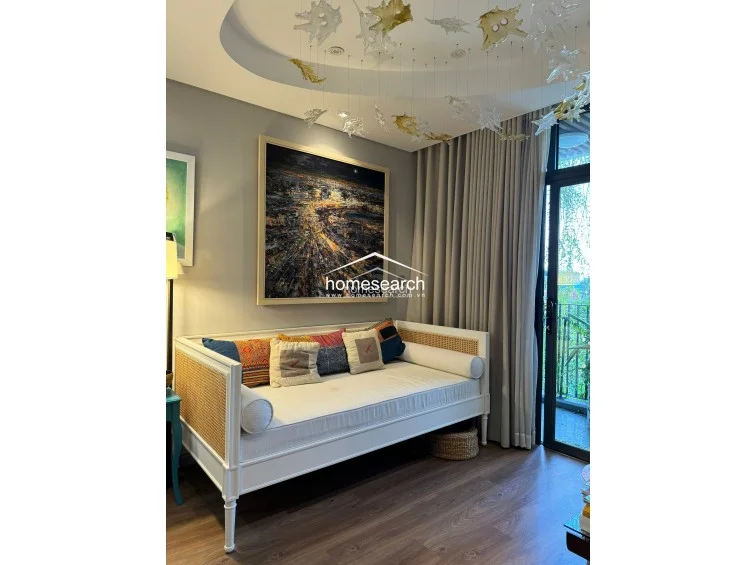 This cozy living space features a stylish daybed with rattan accents, set against a soft grey wall adorned with a large, vibrant cityscape artwork. The room is illuminated by a unique, artistic glass ceiling fixture and enjoys natural light through a floor-to-ceiling glass door that leads to an outdoor area. The flooring consists of warm-toned wood planks, creating a sophisticated and comfortable atmosphere.