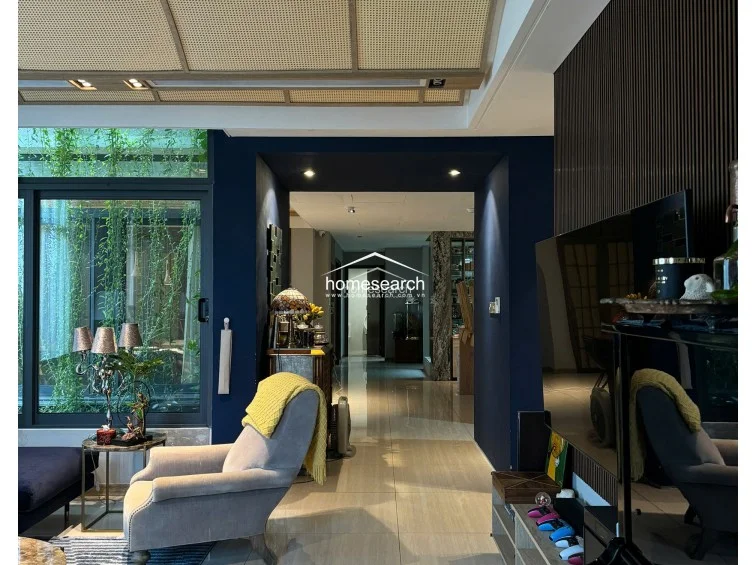 This living area showcases a bold, dark blue accent wall contrasted with a textured, slatted wood panel behind the television. The space flows into a hallway with polished light-colored tile flooring, while a large glass wall provides a lush view of hanging greenery outside. The ceiling features decorative rattan paneling, adding a unique architectural touch to the refined interior.