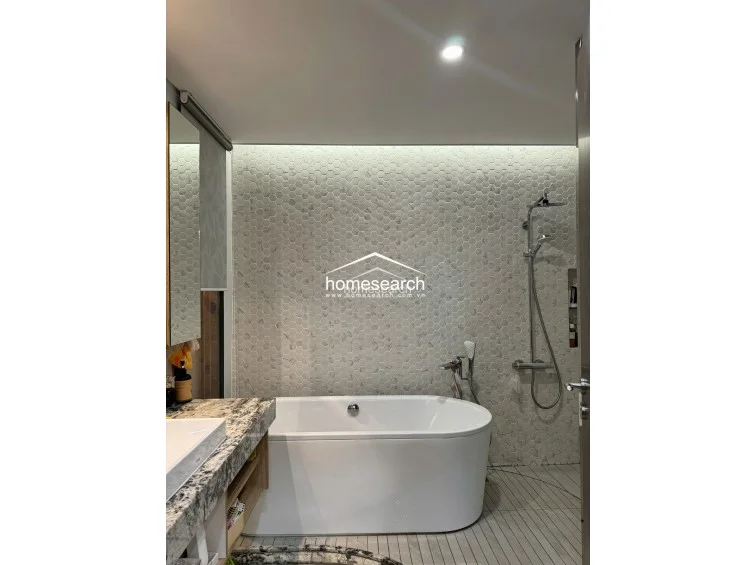 This modern bathroom features a sleek, freestanding white bathtub set against a striking hexagonal marble-tiled feature wall. The vanity is topped with high-end, heavily veined marble, and the space is finished with light-colored floor tiles and recessed ceiling lighting. The overall condition is excellent, reflecting a high-end, contemporary design aesthetic.