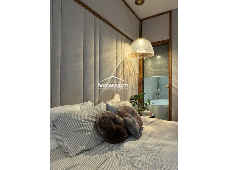 This serene bedroom is defined by a tall, upholstered floor-to-ceiling headboard with vertical channel tufting and warm wood trim. A modern, textured glass pendant light casts soft, ambient shadows, while a glimpse through the side glass panel reveals a private bathroom with a soaking tub. The room maintains a cohesive, high-end design with a neutral color palette and premium finishes.