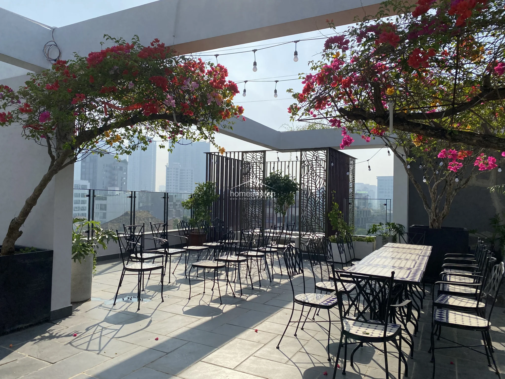This image captures the same rooftop terrace from a slightly different angle, emphasizing the airy, spacious layout and the integration of natural greenery. The stone-tiled flooring and decorative metal privacy screens provide a sophisticated, contemporary aesthetic for outdoor entertaining.