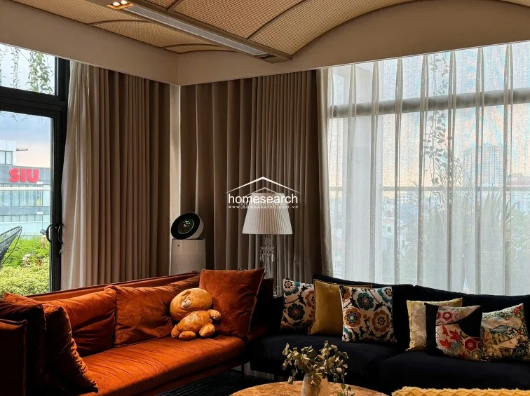 This cozy living room features a striking contrast between a vibrant orange velvet sofa and deep navy blue seating, accented by decorative floral pillows. The room is bathed in natural light from floor-to-ceiling windows draped in elegant, layered curtains, and features a unique vaulted ceiling with woven detailing.