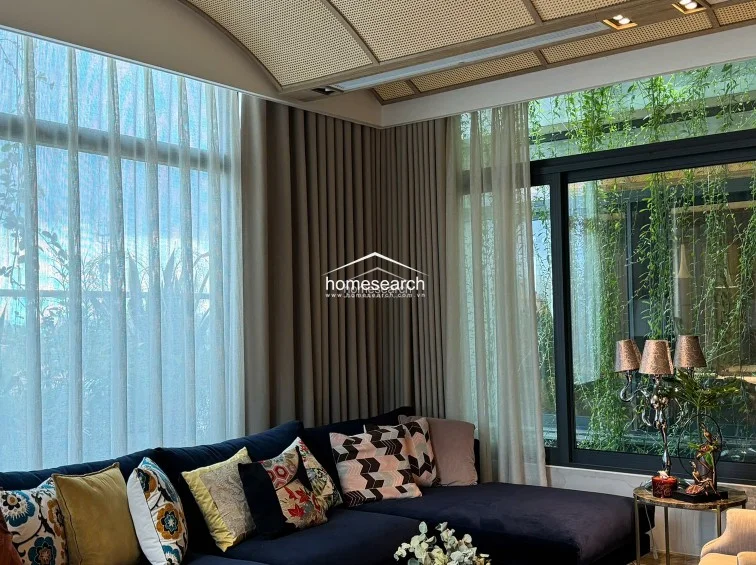 This living area features a sophisticated, modern design with floor-to-ceiling windows that offer a lush, green view, creating a seamless connection to the outdoors. The space is highlighted by a deep navy sectional sofa, a textured rattan-style ceiling detail, and floor-to-ceiling curtains that enhance the room's elegant and well-maintained aesthetic.