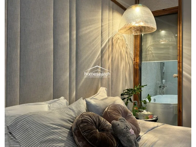 This cozy lounge or bedroom area features a stylish daybed with rattan accents, set against a neutral grey wall adorned with a large, vibrant cityscape artwork. Full-height floor-to-ceiling curtains frame a sliding glass door that provides direct access to a balcony, allowing for ample natural light and a view of lush greenery.