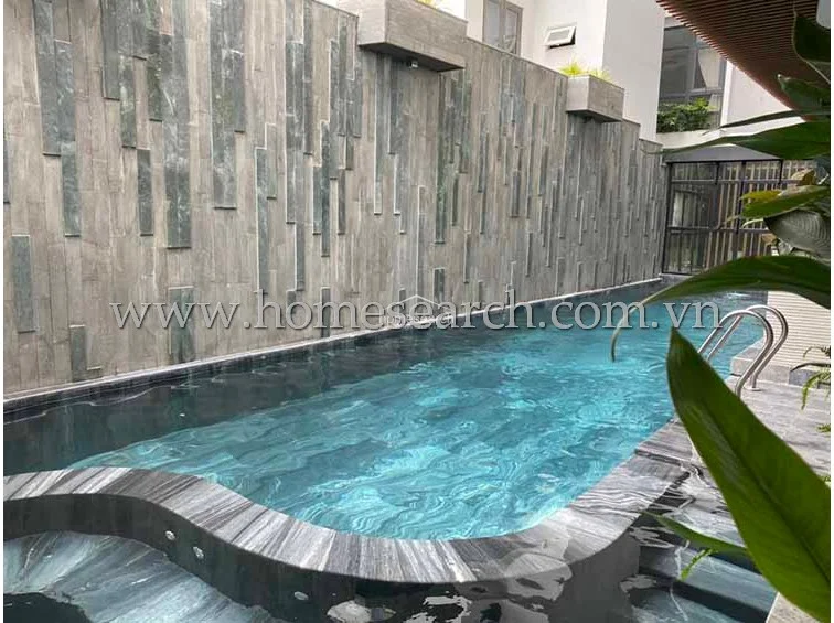 This private outdoor swimming pool is framed by a dramatic, multi-textured stone feature wall that adds significant architectural interest. The pool deck is finished in dark, sophisticated stone, and the water is clear, indicating well-maintained facilities. Lush greenery and a covered patio area suggest a peaceful, resort-style environment.