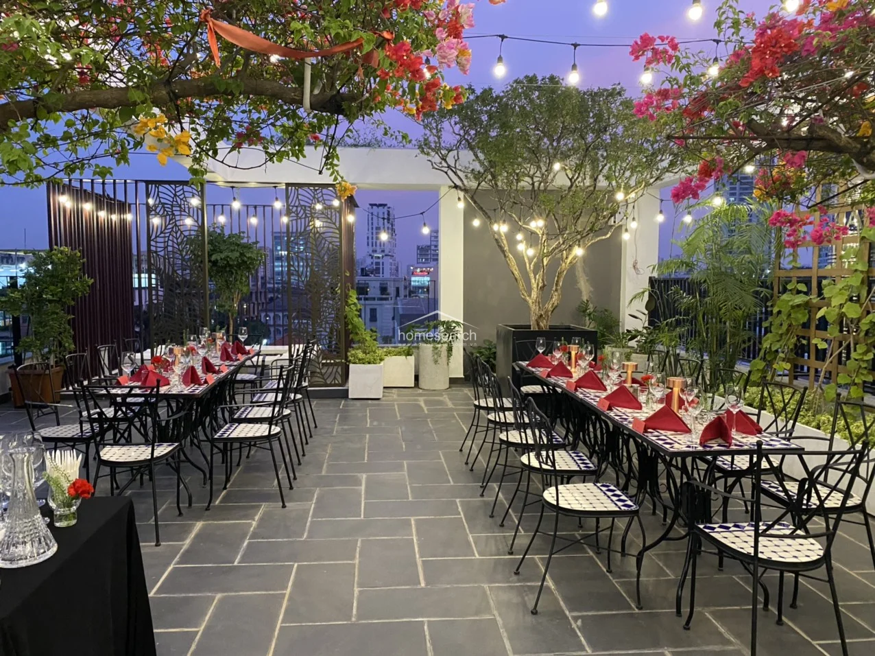 This rooftop terrace is set up for a formal event, featuring multiple dining tables adorned with red napkins and glassware under a canopy of flowering trees. The transition from daylight to dusk highlights the warm glow of the string lights against the backdrop of the surrounding urban architecture.