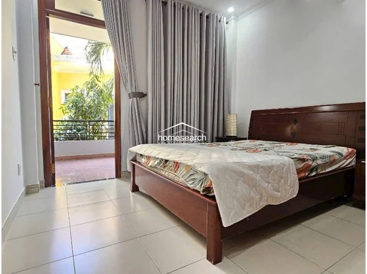 This bedroom features a classic wooden bed frame and a large, built-in wooden wardrobe that matches the room's traditional aesthetic. A floor-to-ceiling glass door with grey curtains provides direct access to a private balcony, allowing for excellent natural light and ventilation.