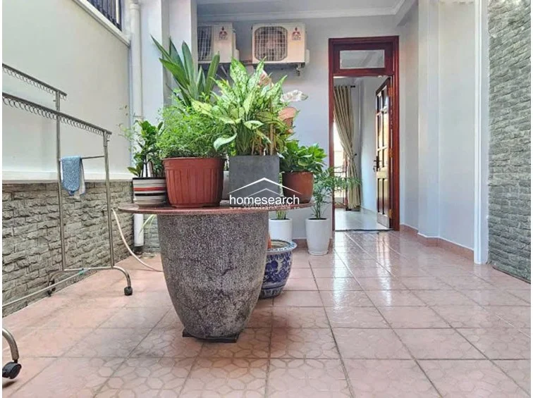 This outdoor balcony or terrace area is finished with light-toned ceramic floor tiles and features a textured stone-cladding wall. The space is currently utilized as a garden nook with various potted plants and includes two external AC compressor units mounted high on the wall.