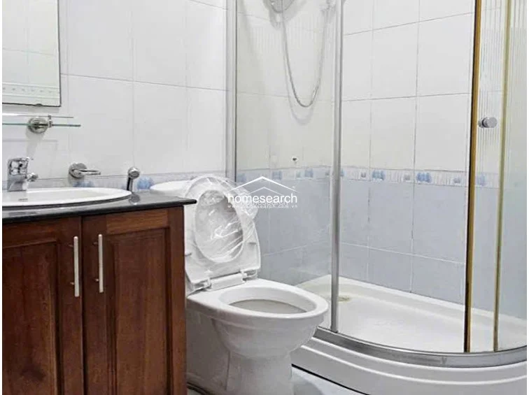 The bathroom is designed with a space-saving corner glass shower enclosure and a wooden vanity cabinet topped with a dark stone counter. It features white wall tiling with a decorative border and appears to be in well-maintained, functional condition.
