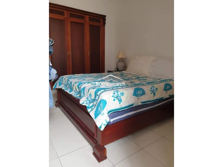 This bedroom is furnished with a sturdy, dark-wood bed frame and a matching full-height wooden wardrobe for integrated storage. The room maintains a neutral color palette with white walls and light-colored floor tiles, creating a simple and functional sleeping environment.