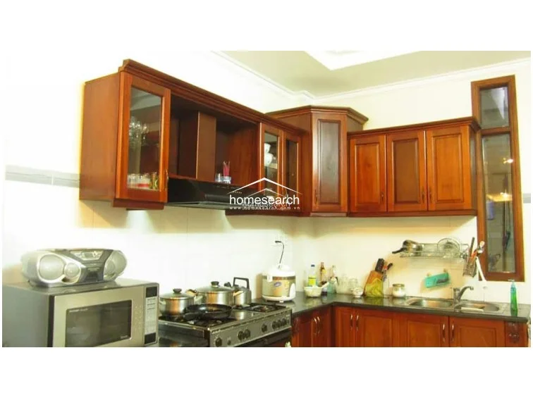This L-shaped kitchen features rich, dark-stained wooden cabinetry that provides ample overhead and base storage. The space is equipped with a modern range hood and a stainless steel sink, set against a backdrop of clean white wall tiles and a dark countertop.