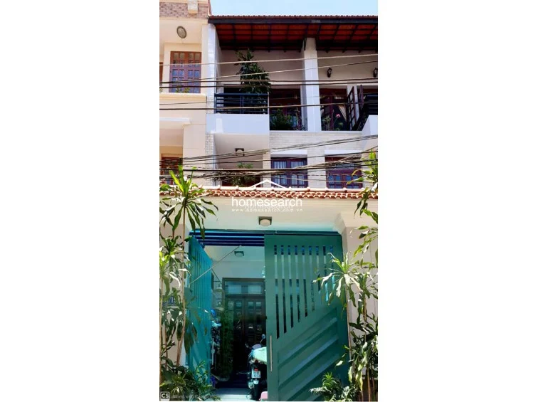 This exterior view shows a multi-story townhouse featuring a green metal gate entrance and balconies on the upper levels. The facade includes dark wood-framed windows, tiled roof detailing, and integrated planters, suggesting a well-maintained urban residence.