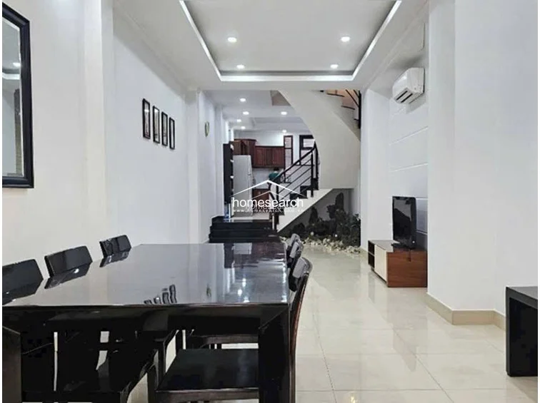 The dining area features a modern, high-gloss dark dining set and polished light-colored tile flooring that extends into the living space. The open-plan layout flows toward a curved staircase and a kitchen area in the back, all illuminated by recessed ceiling lights and a wall-mounted AC unit.