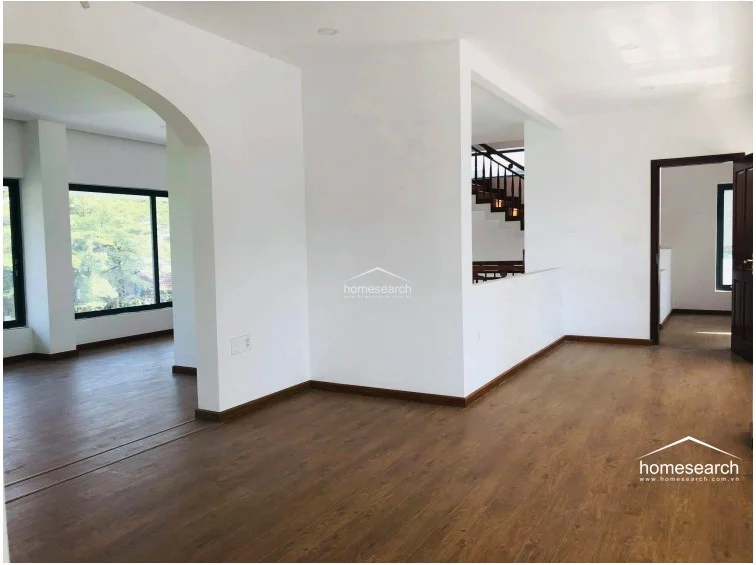 This spacious interior features warm wood-look flooring and clean white walls, connected by elegant, wide archways that promote an open and fluid layout. The space includes a clear view of the wooden staircase and provides easy access to adjacent rooms, maintaining a cohesive and well-lit aesthetic throughout.