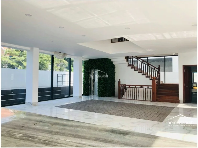 This expansive open-plan living area features a sophisticated mix of white marble flooring accented by a central dark wood inlay, creating a modern and airy atmosphere. The space is anchored by a striking wooden staircase with traditional railings and a unique vertical green wall feature, while floor-to-ceiling glass walls provide abundant natural light and seamless views of the outdoor terrace. The minimalist white ceiling with recessed lighting enhances the sense of volume, and the overall condition appears pristine and well-maintained.