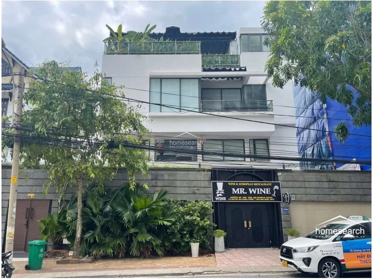 This modern multi-story property features a clean, white minimalist facade with large glass windows and balconies on each level. The ground floor is currently occupied by a commercial establishment, while the upper levels provide private residential or office space, all set behind a stone-walled perimeter with lush greenery.