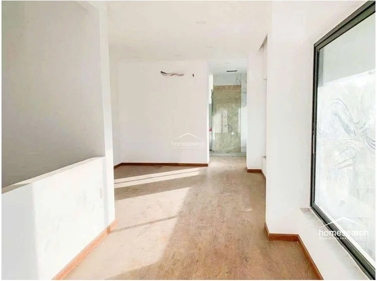 This bright, open-plan room features light-toned wood flooring and crisp white walls, creating a clean and versatile living space. A large floor-to-ceiling window allows for significant natural light, while an open doorway leads directly into an en-suite bathroom with marble-style finishes.