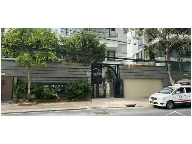 The property exterior showcases a modern, minimalist facade with dark grey stone cladding and a sleek black metal gate. The entrance is framed by lush greenery and mature trees, providing a private and well-maintained street presence.