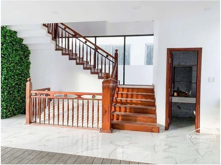 This interior space is defined by a striking wooden staircase with classic balustrades that serves as a central architectural feature. The flooring consists of polished white marble with subtle grey veining, and an adjacent wall features a lush vertical garden, providing a sophisticated contrast between natural elements and modern finishes.
