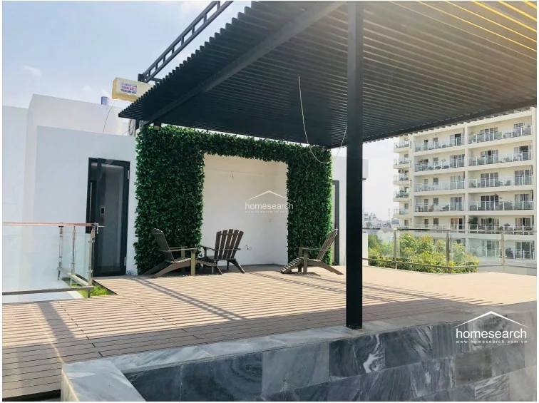 This expansive rooftop terrace offers an ideal outdoor living space, featuring composite decking and a large black metal pergola for shade. A decorative green wall adds a vibrant, natural touch, while the open layout provides a perfect setting for relaxation with views of neighboring buildings.