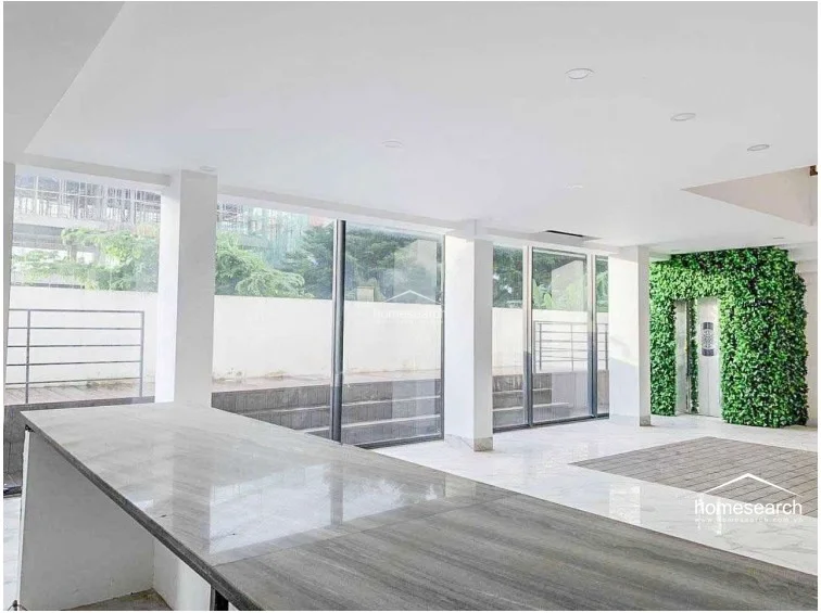 This bright, open-plan living or dining area is defined by high ceilings and large floor-to-ceiling windows that flood the space with natural light. The room features a sleek marble-topped island or counter and a unique green wall feature, creating a modern, airy atmosphere with a seamless connection to the outdoor patio.