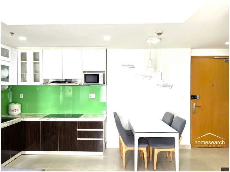 The dining area is situated immediately adjacent to the kitchen, creating an open-concept flow that leads toward the main entrance. The space is illuminated by a modern pendant light fixture above the table and features neutral wall tones accented by decorative wall decals.