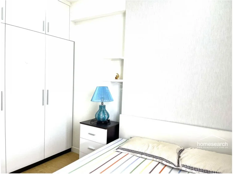 This bedroom view highlights a floor-to-ceiling white wardrobe that maximizes vertical storage space. The room is accented by a decorative blue lamp on a two-tone nightstand, set against a textured white wall finish that adds subtle depth to the space.