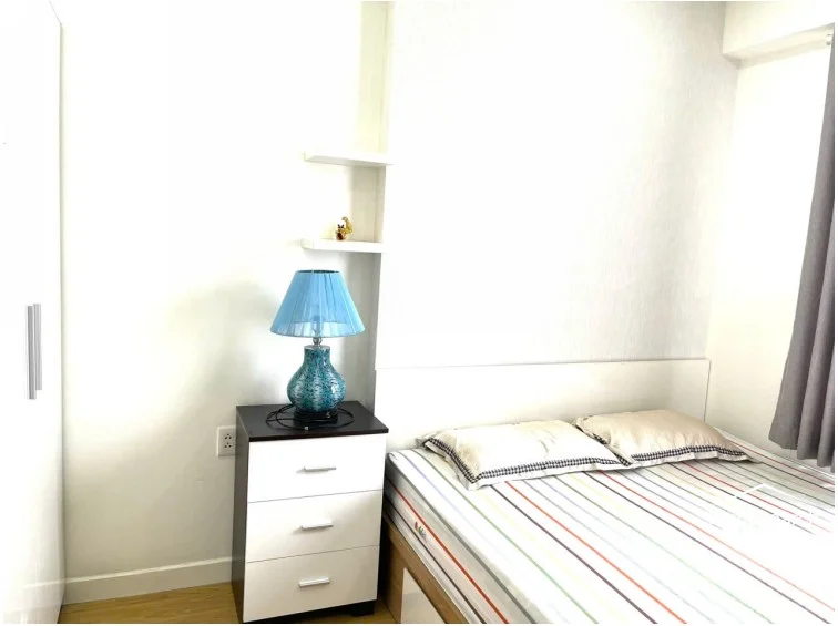 This close-up of the bedroom corner shows a three-drawer nightstand and floating wall shelves, providing efficient storage in a limited footprint. The room is decorated with a vibrant blue lamp, and the overall condition of the finishes and furniture appears excellent.