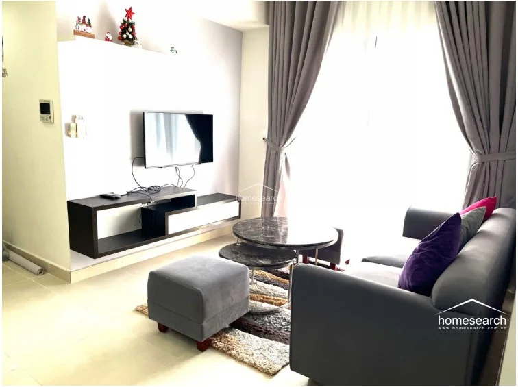 This living room view highlights a cozy seating area with a grey sofa, matching ottoman, and a nested marble coffee table set. Large floor-to-ceiling windows are dressed with heavy grey curtains, allowing for ample natural light, while the wall features decorative holiday accents. The space maintains a cohesive, minimalist aesthetic with neutral tones and clean lines.